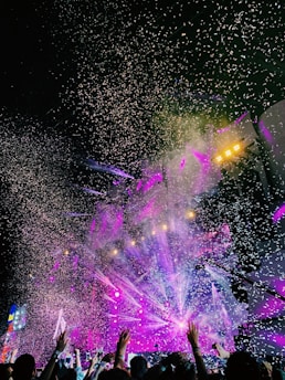 A vibrant concert scene with colorful stage lights and confetti filling the air, creating a dynamic and festive atmosphere. Hands are raised in the crowd, capturing the excitement of the moment.