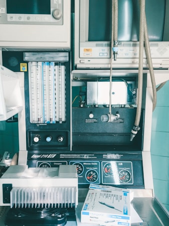 An assortment of medical equipment including monitors, dials, and gauges is present, possibly used for anesthesia or patient monitoring in a healthcare setting. There are various knobs and labels indicating settings for oxygen and nitrous oxide management. A tray contains a packaged medical device labeled 'Hand Piece.'