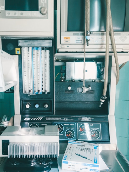 An assortment of medical equipment including monitors, dials, and gauges is present, possibly used for anesthesia or patient monitoring in a healthcare setting. There are various knobs and labels indicating settings for oxygen and nitrous oxide management. A tray contains a packaged medical device labeled 'Hand Piece.'