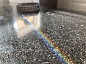 A polished surface, likely a table or countertop, reflects a band of rainbow colors, stretching diagonally across the image. In the background, a blurry object, possibly a cylindrical container or ring, adds a sense of depth.