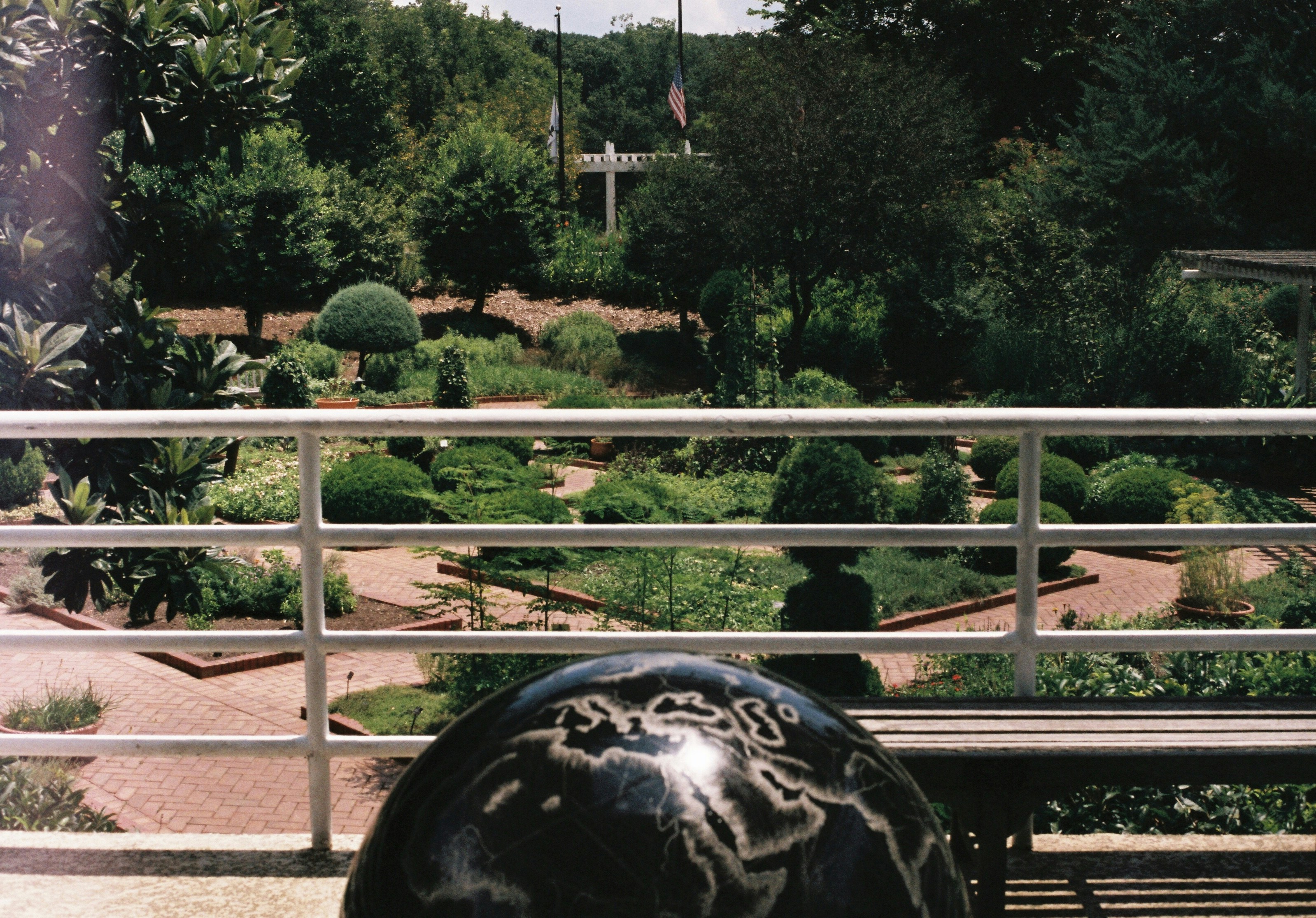 A well-maintained garden with neatly trimmed hedges and various shrubs is visible beyond a white railing. In the foreground, there is a black globe with geographical markings. The setting is lush with greenery and includes brick pathways. Two flagpoles and a white structure are seen in the background surrounded by trees.