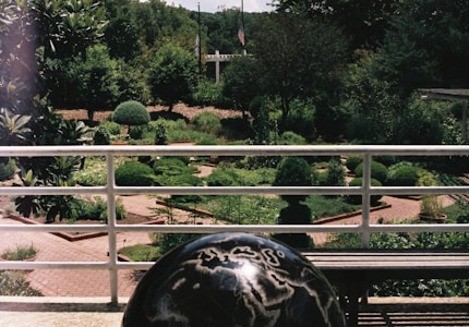 A well-maintained garden with neatly trimmed hedges and various shrubs is visible beyond a white railing. In the foreground, there is a black globe with geographical markings. The setting is lush with greenery and includes brick pathways. Two flagpoles and a white structure are seen in the background surrounded by trees.
