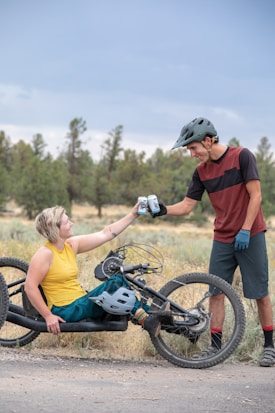 A person sitting on a three-wheel recumbent bike extends a drink to another person standing nearby. Both are wearing bike helmets and casual biking attire. They are in an outdoor setting with trees in the background and appear to be enjoying a break.