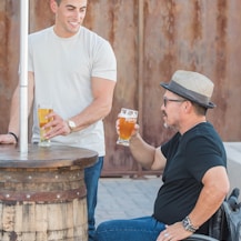 man sitting on wheelchair holding near full clear drinking glass beside standing and smiling man