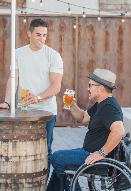 man sitting on wheelchair holding near full clear drinking glass beside standing and smiling man
