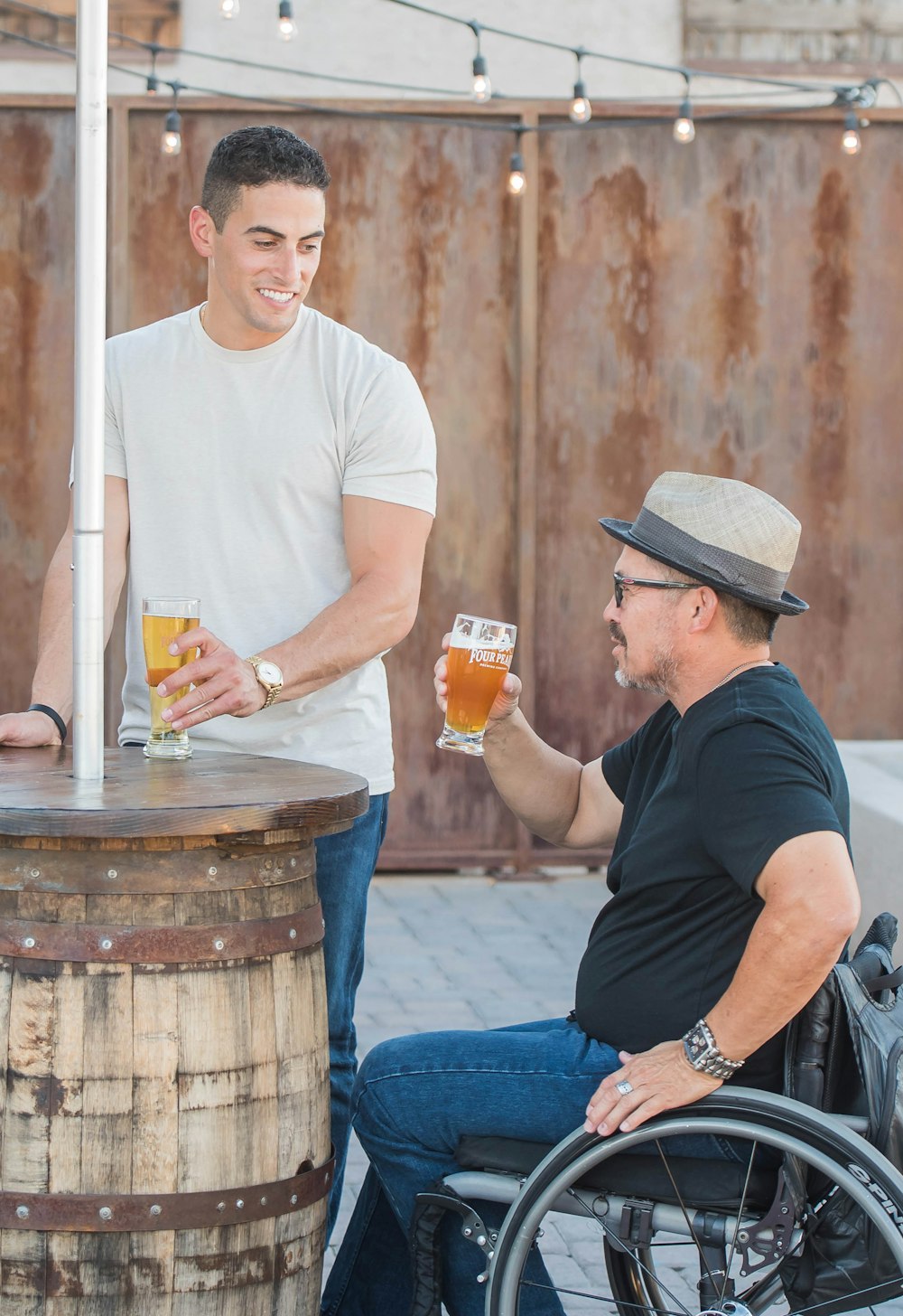 man sitting on wheelchair holding near full clear drinking glass beside standing and smiling man