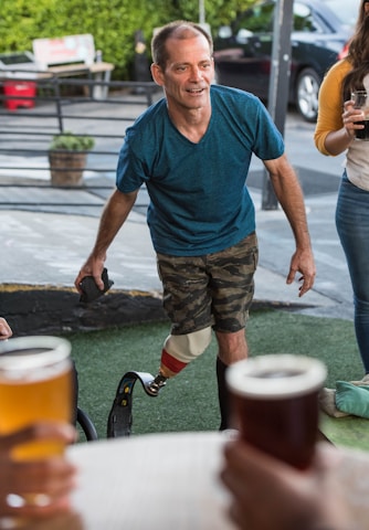 A man wearing a blue shirt and camouflage shorts with a prosthetic leg appears to be interacting amiably in an outdoor setting. He is surrounded by a social group, with hands holding beer glasses visible in the foreground. The setting seems relaxed, with potted plants and parked cars seen in the background.