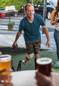A man wearing a blue shirt and camouflage shorts with a prosthetic leg appears to be interacting amiably in an outdoor setting. He is surrounded by a social group, with hands holding beer glasses visible in the foreground. The setting seems relaxed, with potted plants and parked cars seen in the background.