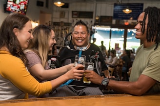 Four people are sitting at a bar, smiling and clinking their glasses together in a celebratory toast. The setting appears to be a casual, well-lit bar or pub with a warm and friendly atmosphere. One person uses a wheelchair and is equipped with adaptive devices to assist with drinking.