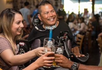 A group of people clinks glasses together in a toast at a lively social gathering. They are smiling and appear to be enjoying each other's company. The setting is a warmly lit indoor space, possibly a bar or restaurant.