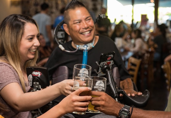 A group of people clinks glasses together in a toast at a lively social gathering. They are smiling and appear to be enjoying each other's company. The setting is a warmly lit indoor space, possibly a bar or restaurant.