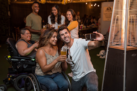A group of smiling people are gathered together in a celebratory atmosphere. Two individuals are in the foreground, with one person taking a selfie while holding a beer. The setting appears to be a lively social event with warm lighting and an outdoor heater nearby.