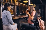 Two women are sitting at a bar counter, each holding a glass of beer. One woman is wearing a denim jacket and skirt, while the other is in a pink shirt and seated in a wheelchair. The bar has a warm, inviting atmosphere with dim lighting and wooden accents. A few people are visible in the background, engaged in their own activities.