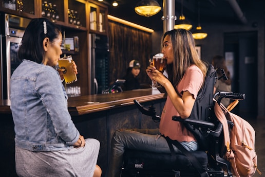 Two women are sitting at a bar counter, each holding a glass of beer. One woman is wearing a denim jacket and skirt, while the other is in a pink shirt and seated in a wheelchair. The bar has a warm, inviting atmosphere with dim lighting and wooden accents. A few people are visible in the background, engaged in their own activities.