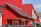 A modern residential building with bright red walls and a matching red roof featuring dark shingles. The structure includes multiple white-framed windows and an entrance door with decorative glass. A portion of a gray fence is visible in the foreground, and the sky is clear and blue.