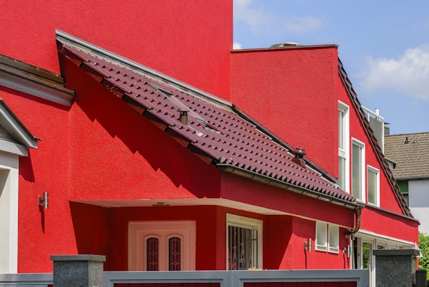 A modern residential building with bright red walls and a matching red roof featuring dark shingles. The structure includes multiple white-framed windows and an entrance door with decorative glass. A portion of a gray fence is visible in the foreground, and the sky is clear and blue.