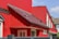 A modern residential building with bright red walls and a matching red roof featuring dark shingles. The structure includes multiple white-framed windows and an entrance door with decorative glass. A portion of a gray fence is visible in the foreground, and the sky is clear and blue.