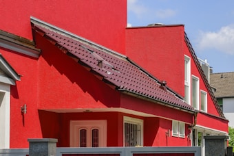 A modern residential building with bright red walls and a matching red roof featuring dark shingles. The structure includes multiple white-framed windows and an entrance door with decorative glass. A portion of a gray fence is visible in the foreground, and the sky is clear and blue.