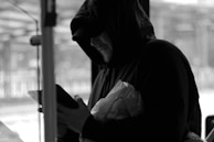 A person wearing a hooded jacket is intently looking at a smartphone or tablet. The individual is partially obscured by shadow, creating a mysterious atmosphere. They are holding a plastic bag, possibly containing groceries or personal items, and are situated in an urban environment, as inferred from the windows and blurred cityscape in the background.