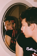 A person with short dark hair is gazing intently at their reflection in a round mirror. Two horizontal red streaks are visible across their black shirt and reflection. The background is softly blurred, drawing focus to the individual's face and expression.