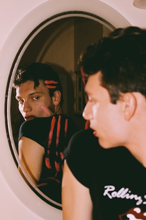 A person with short dark hair is gazing intently at their reflection in a round mirror. Two horizontal red streaks are visible across their black shirt and reflection. The background is softly blurred, drawing focus to the individual's face and expression.
