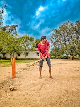 A person is standing outdoors on a dirt surface, holding a cricket bat and preparing to play. The setting includes stumps positioned nearby, and the backdrop consists of lush green trees and a building partially obscured by foliage. The sky is overcast with dramatic dark blue clouds.