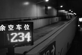 A black and white image of an uphill parking garage entrance with a digital sign displaying the number '234'. Overhead lights illuminate the path, and there are some Asian characters indicating available parking spaces.