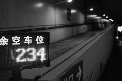 A black and white image of an uphill parking garage entrance with a digital sign displaying the number '234'. Overhead lights illuminate the path, and there are some Asian characters indicating available parking spaces.