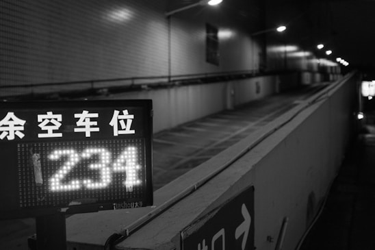 A black and white image of an uphill parking garage entrance with a digital sign displaying the number '234'. Overhead lights illuminate the path, and there are some Asian characters indicating available parking spaces.