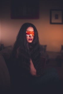 A woman with long, dark hair sits on a sofa in a dimly lit room. She has a colorful blindfold covering her eyes, and her lips are painted red. The background features blurred frames and a warm ambiance.
