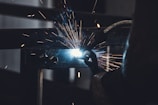 Sparks fly as a person welds a piece of metal. The bright light of the welding arc illuminates the surrounding area in contrast with the dark background. Industrial machinery and tools are visible, suggesting a workshop or manufacturing environment.