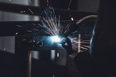 Sparks fly as a person welds a piece of metal. The bright light of the welding arc illuminates the surrounding area in contrast with the dark background. Industrial machinery and tools are visible, suggesting a workshop or manufacturing environment.
