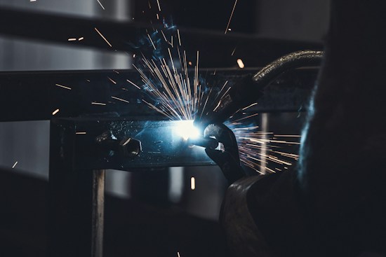 Sparks fly as a person welds a piece of metal. The bright light of the welding arc illuminates the surrounding area in contrast with the dark background. Industrial machinery and tools are visible, suggesting a workshop or manufacturing environment.