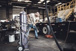 An older man is working in a garage or workshop environment, using tools on a trailer. Surroundings include various equipment and materials such as a large gas cylinder and a hydraulic jack. The space is industrial with stairs leading to a loft and shelves lined with parts.