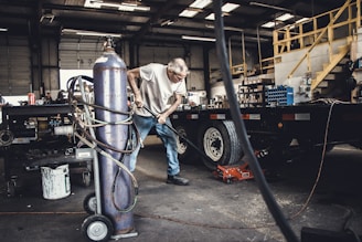 An older man is working in a garage or workshop environment, using tools on a trailer. Surroundings include various equipment and materials such as a large gas cylinder and a hydraulic jack. The space is industrial with stairs leading to a loft and shelves lined with parts.