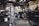 An older man is working in a garage or workshop environment, using tools on a trailer. Surroundings include various equipment and materials such as a large gas cylinder and a hydraulic jack. The space is industrial with stairs leading to a loft and shelves lined with parts.