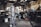 An older man is working in a garage or workshop environment, using tools on a trailer. Surroundings include various equipment and materials such as a large gas cylinder and a hydraulic jack. The space is industrial with stairs leading to a loft and shelves lined with parts.