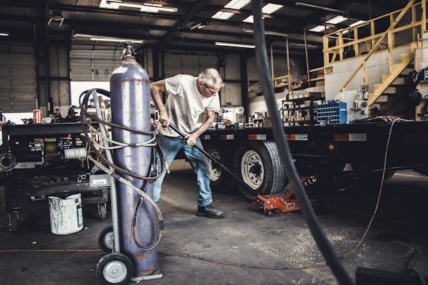 An older man is working in a garage or workshop environment, using tools on a trailer. Surroundings include various equipment and materials such as a large gas cylinder and a hydraulic jack. The space is industrial with stairs leading to a loft and shelves lined with parts.
