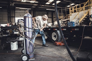 An older man is working in a garage or workshop environment, using tools on a trailer. Surroundings include various equipment and materials such as a large gas cylinder and a hydraulic jack. The space is industrial with stairs leading to a loft and shelves lined with parts.