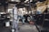 An older man is working in a garage or workshop environment, using tools on a trailer. Surroundings include various equipment and materials such as a large gas cylinder and a hydraulic jack. The space is industrial with stairs leading to a loft and shelves lined with parts.