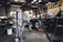 An older man is working in a garage or workshop environment, using tools on a trailer. Surroundings include various equipment and materials such as a large gas cylinder and a hydraulic jack. The space is industrial with stairs leading to a loft and shelves lined with parts.