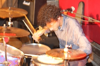 A person with curly hair is playing drums, surrounded by a microphone and cymbals. The setting appears to be a music practice space with red panels in the background. The motion blur suggests dynamic movement as they strike the drums with sticks.