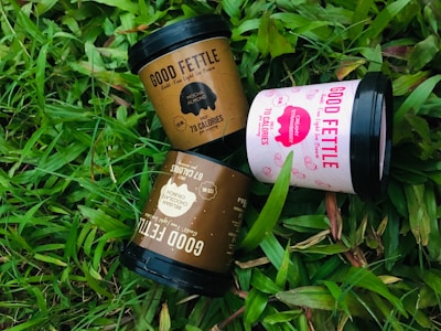 Three ice cream containers labeled 'Good Fettle' are placed on vibrant green grass. The containers include flavors such as Mocha Almond, Creamy Strawberry, and Belgian Chocolate Crunch. Each container features calorie information and playful design elements.