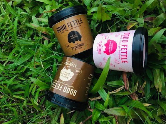Three ice cream containers labeled 'Good Fettle' are placed on vibrant green grass. The containers include flavors such as Mocha Almond, Creamy Strawberry, and Belgian Chocolate Crunch. Each container features calorie information and playful design elements.