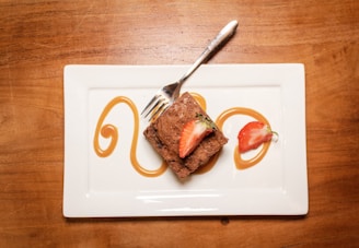 A square chocolate brownie is elegantly placed on a white rectangular plate. It is garnished with two slices of fresh strawberries on top. Artistic caramel swirls decorate the plate around the brownie. A shiny fork rests on the plate, suggesting the dessert is ready to be eaten. The table underneath is wooden, complementing the dessert’s presentation.