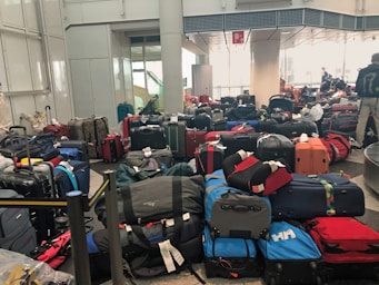 A crowded airport baggage claim area filled with numerous suitcases of various colors and sizes, placed haphazardly on the floor. In the background, there are a few people, some of whom seem to be searching for their luggage.