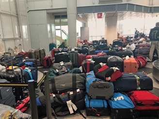 A crowded airport baggage claim area filled with numerous suitcases of various colors and sizes, placed haphazardly on the floor. In the background, there are a few people, some of whom seem to be searching for their luggage.