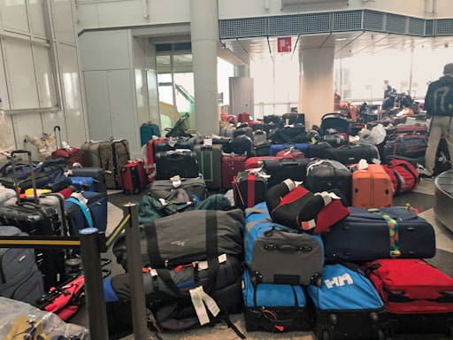 A crowded airport baggage claim area filled with numerous suitcases of various colors and sizes, placed haphazardly on the floor. In the background, there are a few people, some of whom seem to be searching for their luggage.