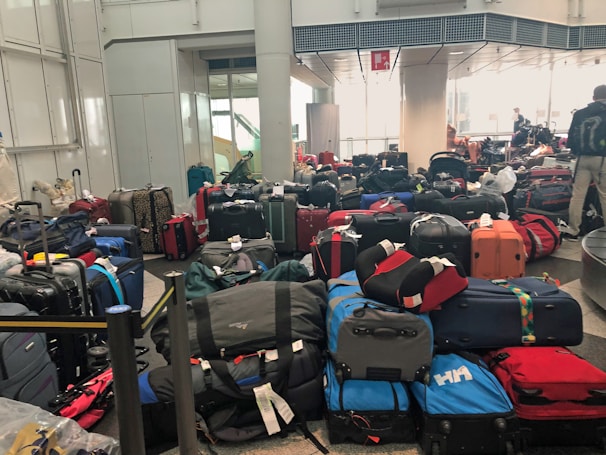 A crowded airport baggage claim area filled with numerous suitcases of various colors and sizes, placed haphazardly on the floor. In the background, there are a few people, some of whom seem to be searching for their luggage.