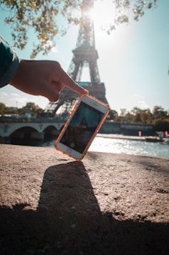 A hand holding a smartphone near the Eiffel Tower under a clear blue sky. The phone is capturing the iconic structure in its camera. Sunlight creates a dynamic play of light and shadow, with a shadow of the phone elongated on the concrete surface. Trees with green leaves surround the background, contributing to the bright, lively atmosphere.